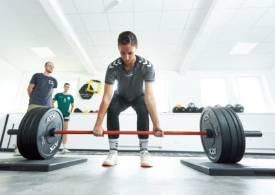 Sportlerin an der Langhantel Blick von unten vorne mit Personal Trainer und Physiotherapeut Malte Klett und weiterer Sportlerin wartend